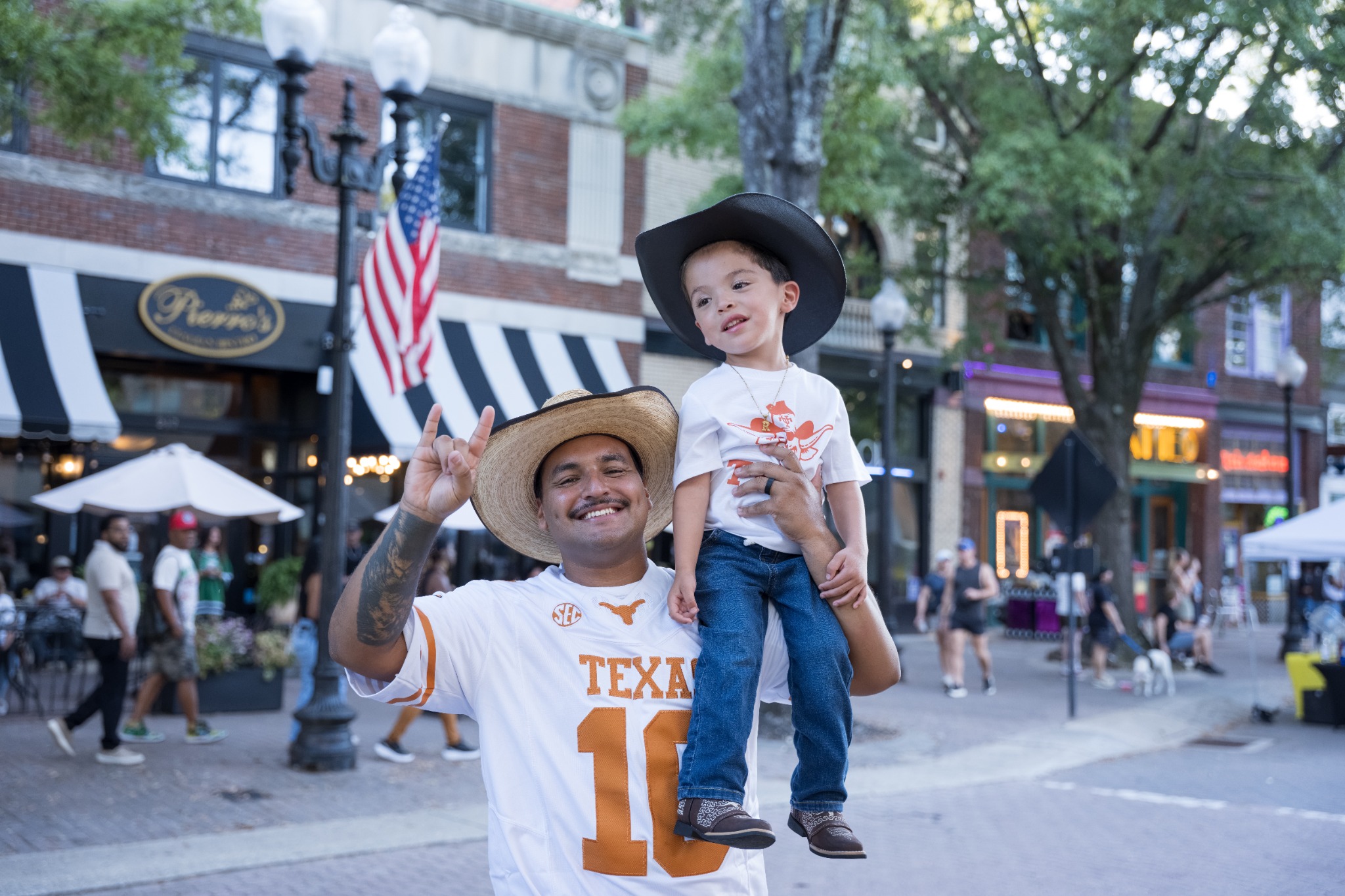 Event guest in a straw cowboy hat holding a child as they look along the street
