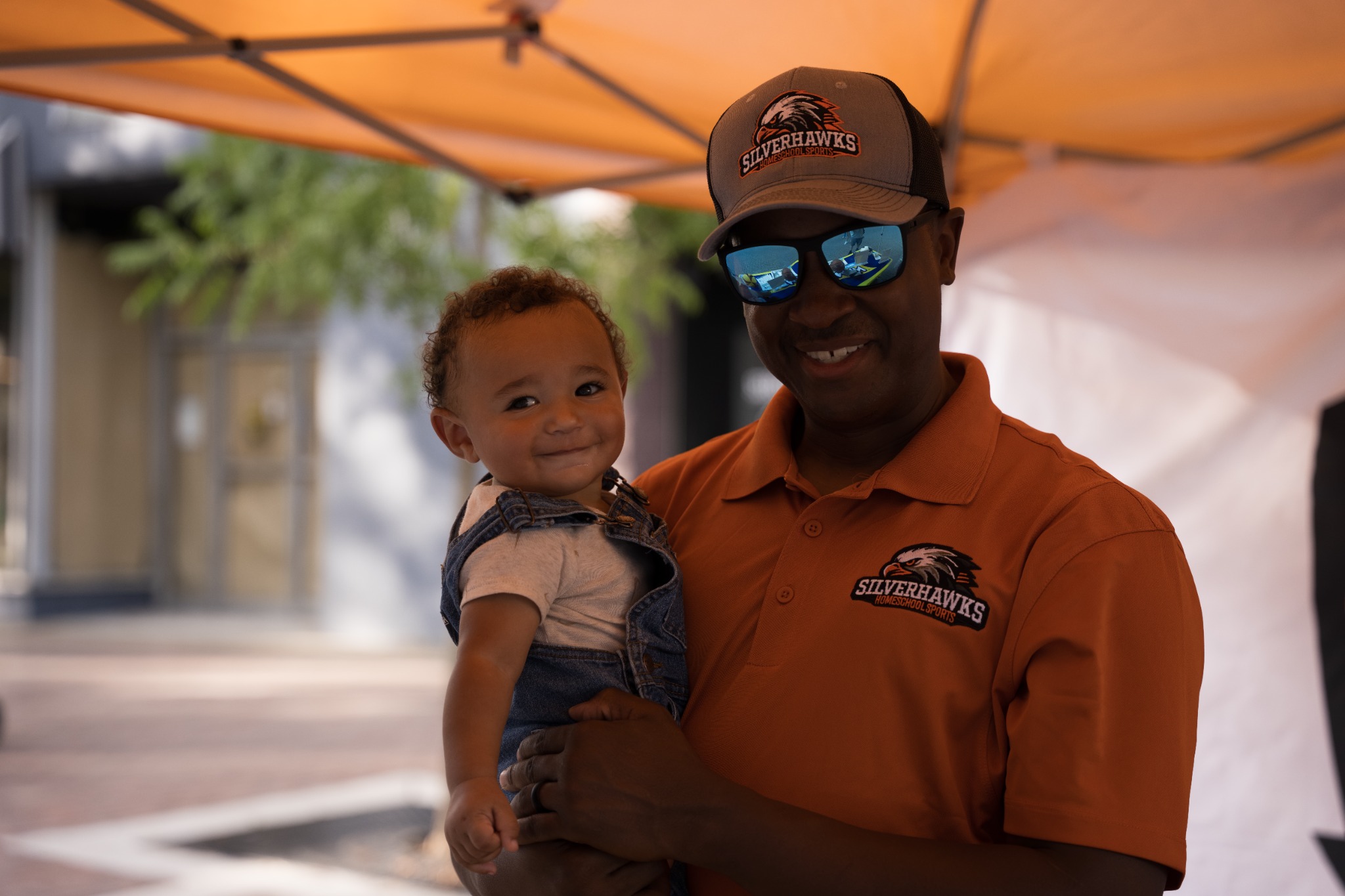 Event guest in an orange polo and blue sunglasses holding a smiling baby under a canopy